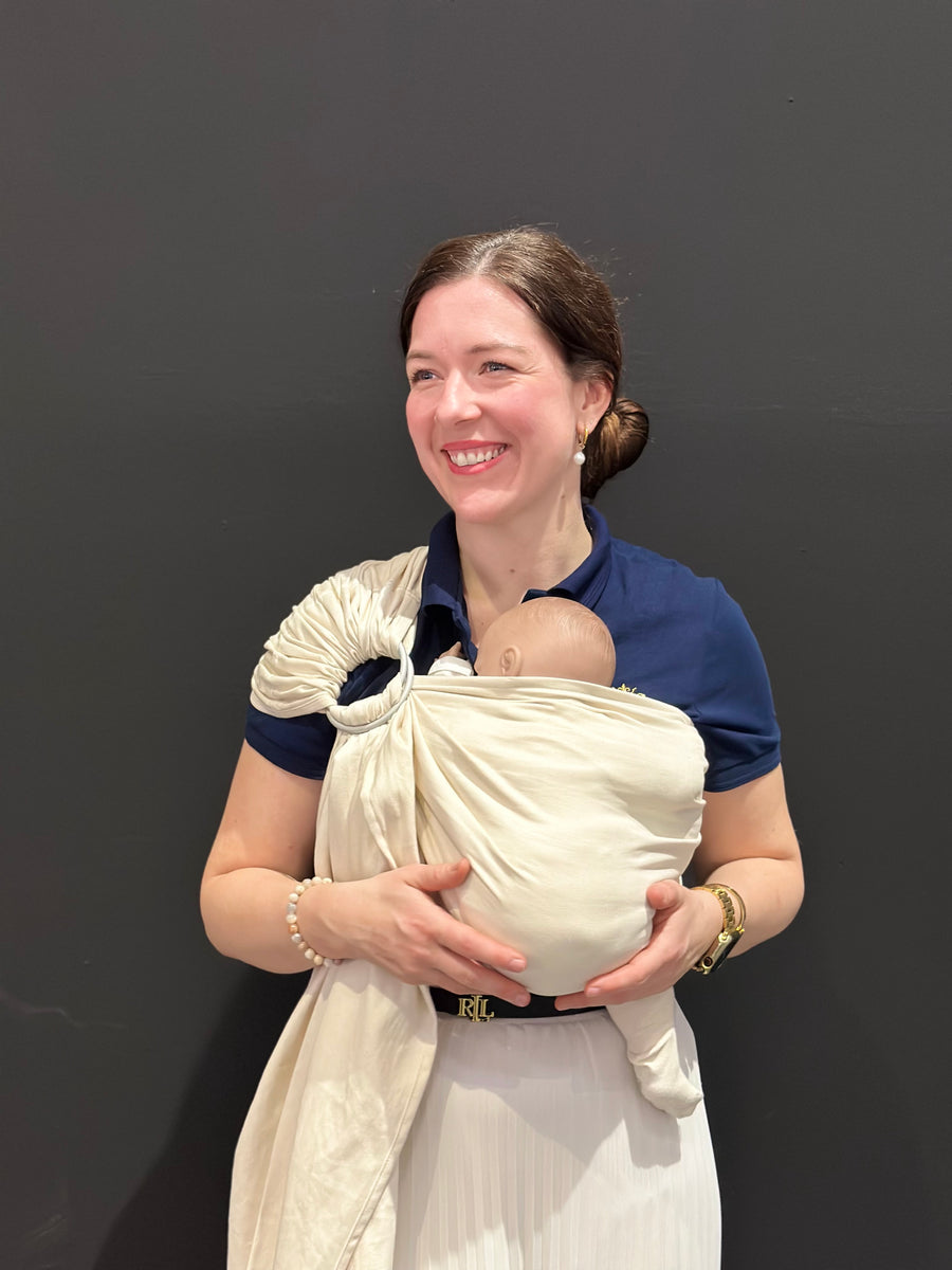 Ringsling Natur Beige: Eine Frau trägt eine Puppe im hellen Tuch vor der Brust, an der Schulter die Ringe.
