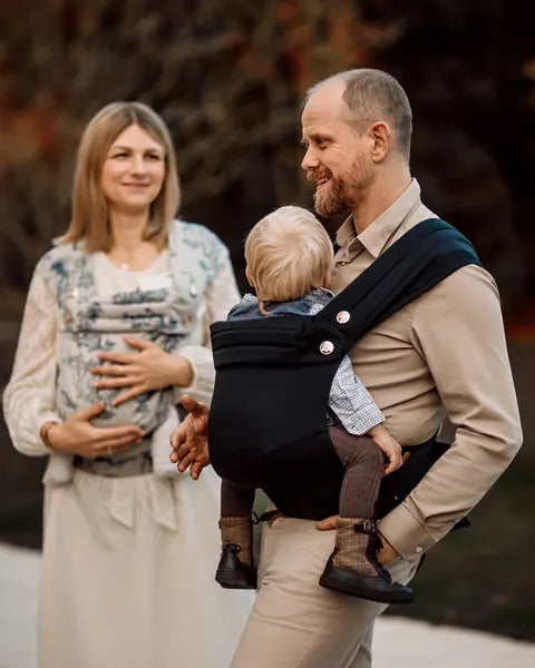 Eine Frau mit der MonDoudou Toile de Jouy und ein Mann mit der MonDoudou Noir tragen je ein Kind, er wendet sich ihr lachend zu.