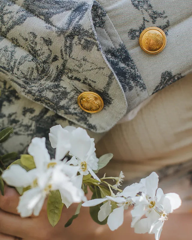 Zwei Goldknöpfe mit Wappenprägung am Träger einer MonDoudou Toile de Jouy, davor ein Zweig mit weißen Blüten.