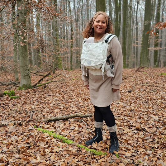 KAAMI Click&Cuddle: Eine Frau mit der hellen Trage vor dem Bauch steht in einem herbstlichen Wald.
