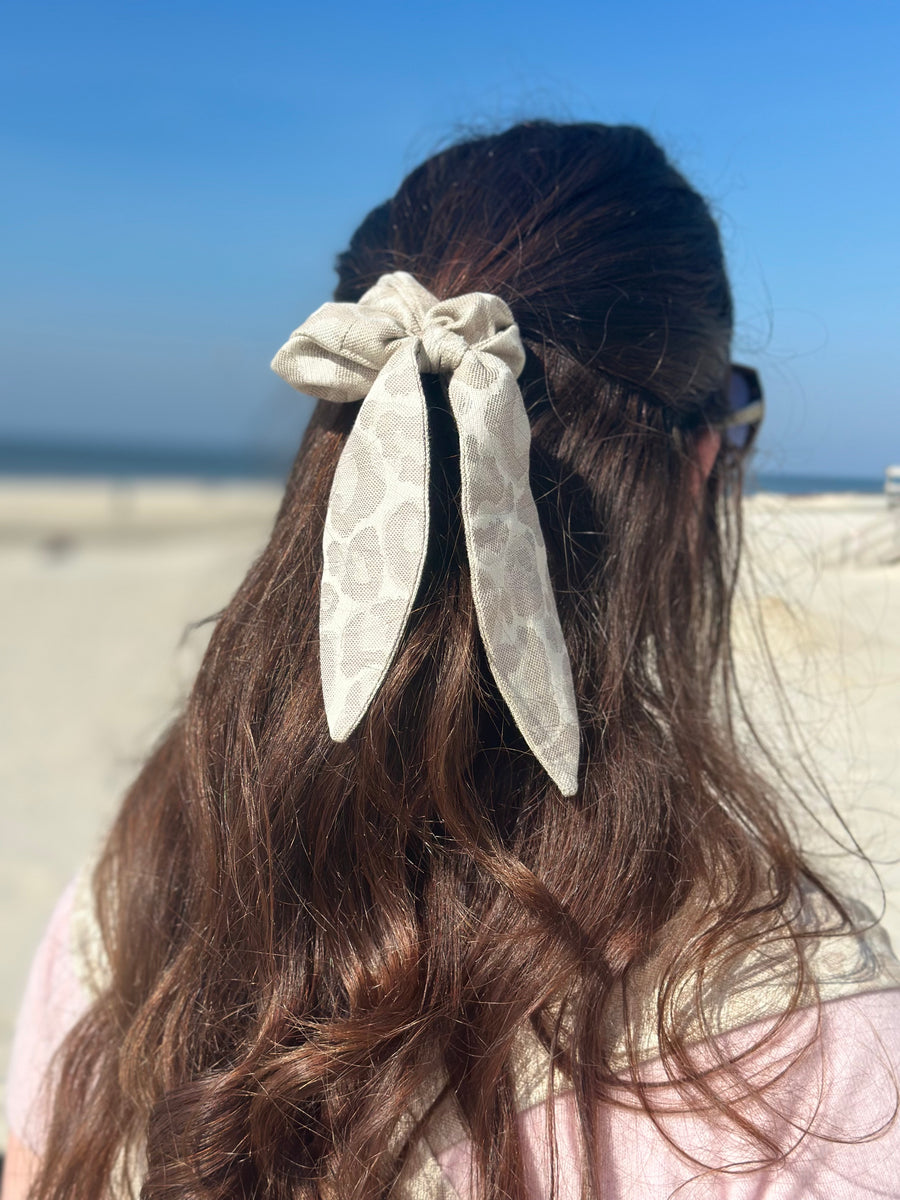 Eine Frau am Strand mit einem Scrunchie mit Schleife im hellen Leopardenmuster im Haar.