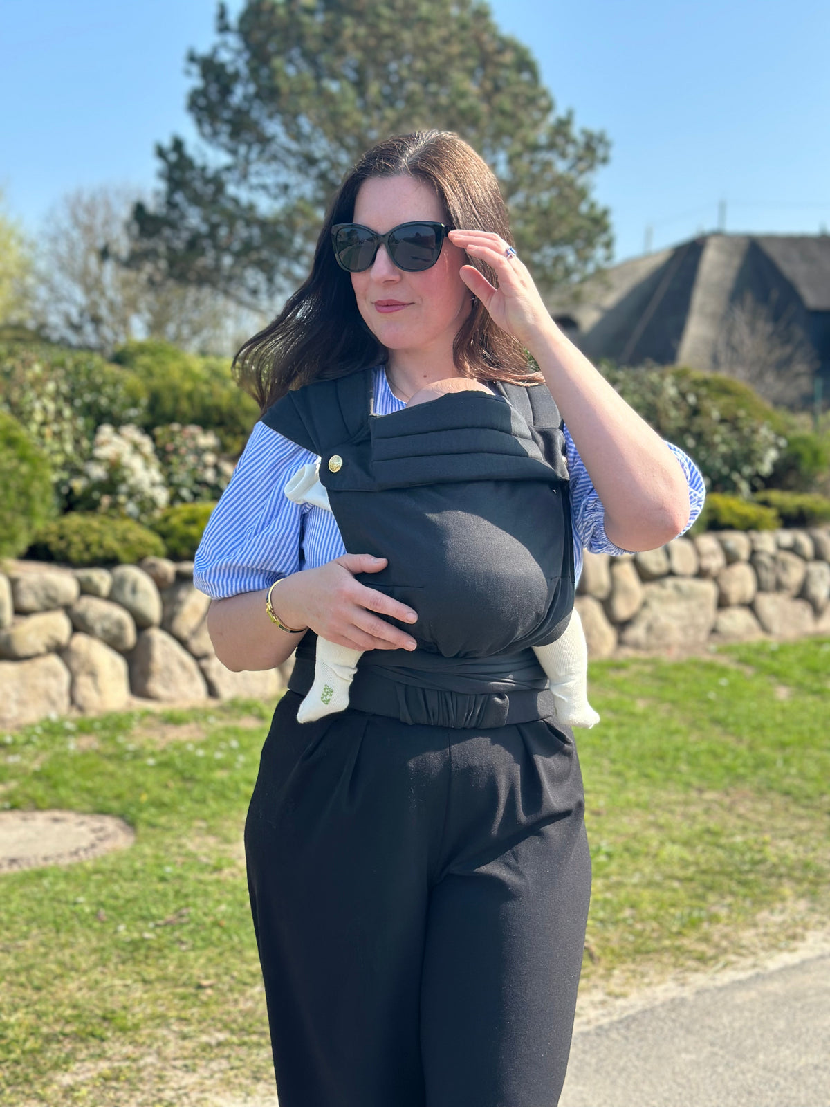 MonDoudou Noir: Eine Frau mit Sonnenbrille trägt ihr Kind vor dem Bauch, hinter ihr ein Steinwall und Reetdachhäuser.