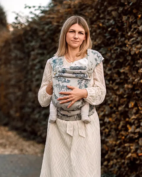 MonDoudou Toile de Jouy: Eine Frau im hellen Kleid trägt ihr Kind vor dem Bauch, hinter ihr eine hohe Hecke.